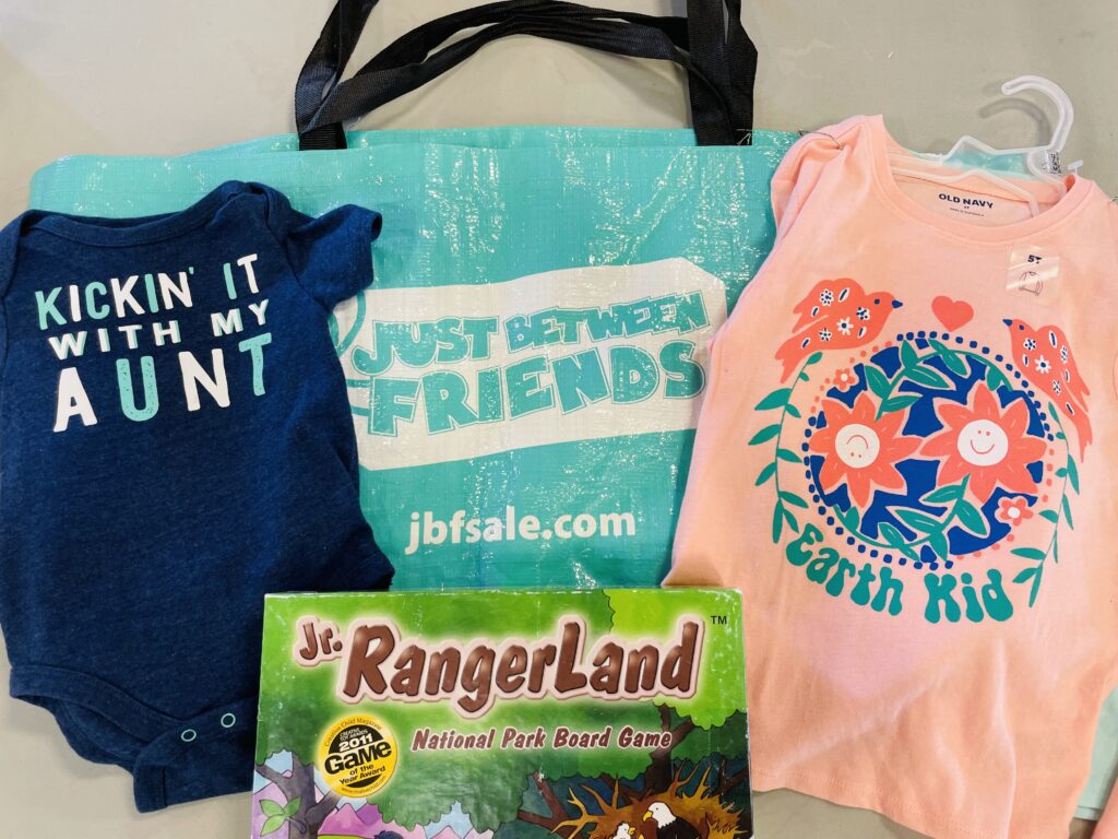 Picture of Just Between Friends Consignment reusable shopping bag with logo and consignment finds which include toddler shirt that says "Earth Kid", a onesie that say "Kickin it with my Aunt" and a Jr Rangerland used game